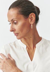 Zlaté slzovité náušnice visia z ucha modelky a kontrastujú s bielym, jednoduchým topom bez goliera. Hladká textura, elegantný design.
