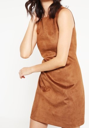 Mujer con vestido sin mangas de ante marrón con detalles sutiles en las costuras, tocándose el cabello e inclinándose ligeramente hacia adelante sobre un fondo blanco.