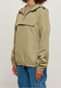 Jasnooliwkowa bluza z kapturem, z zamkiem z przodu, elastycznym paskiem w talii i szerokimi mankietami. Posiada kieszeń na piersi z klapką.