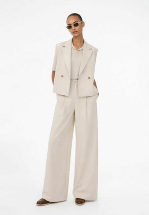 Kvinde iført beige bukser med brede ben, matchende ærmeløs blazer, poloshirt, brune sko og smalle solbriller, stående med hænderne i lommerne.