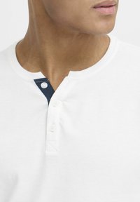 Cămașă henley din bumbac alb, cu guler rotund, având un placket cu nasturi navy și două nasturi, textură netedă și design simplu.