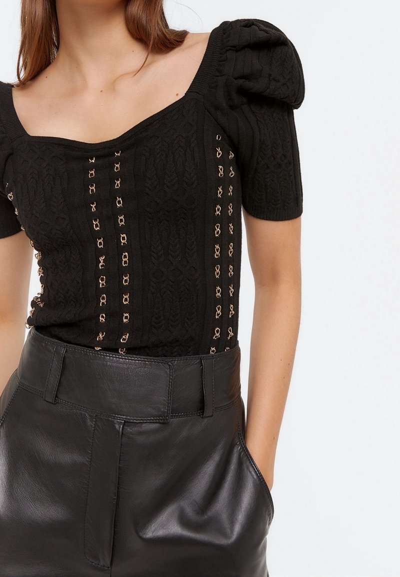 Top nero lavorato a maglia con scollatura quadrata, maniche a sbuffo, motivo testurizzato e dettagli dorati. Abbinato a pantaloni shorts in pelle nera a vita alta.