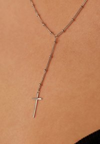 Zilveren kruis hanger ketting met een delicate ketting en kleine kralen, hangend tegen een huidtint achtergrond.