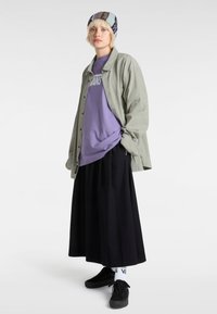 Giacca in corda verde chiaro oversized sopra una maglietta viola, gonna lunga nera, sneakers nere e berretto con motivo. Calze bianche con logo.