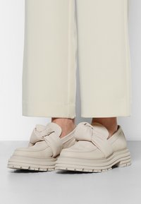 Beiga slip-on-skor med en knuten detalj, med en tjock, strukturerad sula och en slät läderovandel. Bärs med krämfärgade vida byxor.