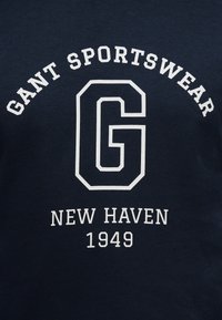 Σκούρο μπλε ύφασμα με λευκό κείμενο: «GANT SPORTSWEAR» κυρτό πάνω από ένα μεγάλο γράμμα «G», με τα «NEW HAVEN 1949» από κάτω.