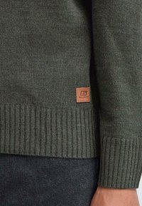 Pulover tricotat de un verde închis, cu margini și manșete ondulate, având o mică etichetă din piele maro pe partea inferioară. Textura este moale și densă.