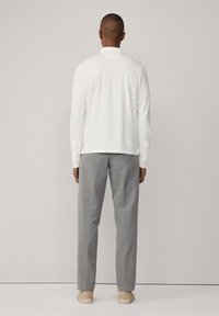 Camicia bianca a maniche lunghe con polsini a bottone, pantaloni grigi su misura e scarpe beige. Modello visto da dietro su uno sfondo semplice.