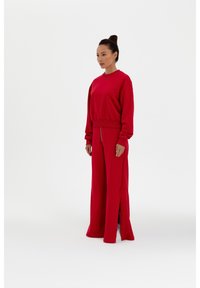 Rotes zweiteiliges Outfit, bestehend aus einem Sweatshirt mit Rundhalsausschnitt und weiten Hosen mit seitlichen Schlitzen. Der Stoff wirkt weich und strukturiert.