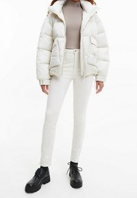 Chaqueta de plumas blanca con cuello alto y cierre de cremallera, que cuenta con dos bolsillos frontales. Combinada con unos jeans blancos ajustados y botas negras con cordones.