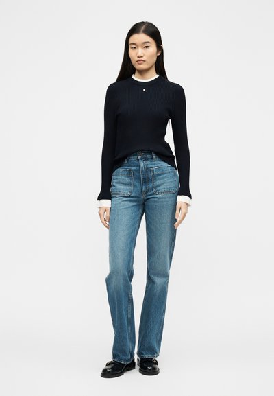 Γυναίκα που στέκεται φορώντας μαύρο ριμπ πλεκτό πουλόβερ με λευκές μανσέτες, τζιν ψηλόμεσο με τσέπες επιδιορθωμένες, και μαύρα loafers σε λευκό φόντο.