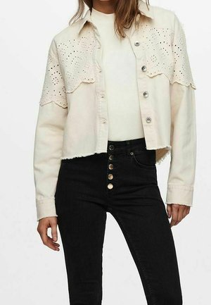 Kvinde iført en cremefarvet cropped jakke med hulmønster detaljer over en hvid top og højtaljede sorte jeans med fem synlige knapper.