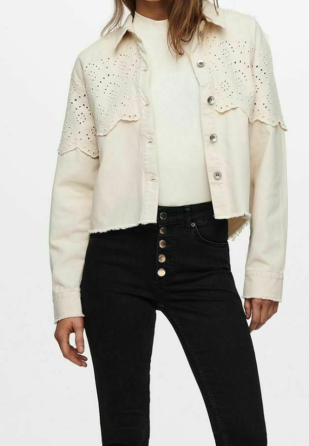 Femme portant une veste courte crème avec des détails œillets par-dessus un top blanc et un jean noir taille haute avec cinq boutons visibles.