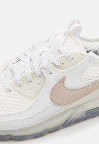 Vit Nike-sneaker med beige swoosh, tjocka vita snören, nätövre och genomskinlig sula med synlig luftdämpning.