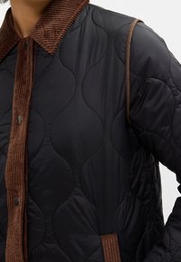Personne portant une veste matelassée noire avec un col en velours côtelé marron, une patte de boutonnage et un bord de poche, montrée de la poitrine à l'épaule.