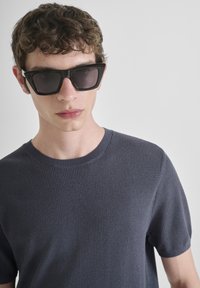 Crne pravokutne sunčane naočale s tamnim staklima. Nosene s teksturiranom tamnoplavom majicom kratkih rukava s okruglim izrezom. Glatka put.