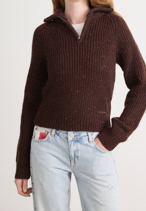 Femme portant un pull en maille côtelée marron foncé avec un col demi-fermeture éclair associé à un jean bleu clair usé.