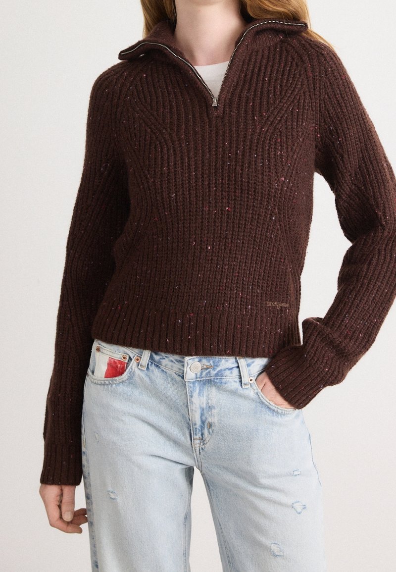 Femme portant un pull en maille côtelée marron foncé avec un col demi-fermeture éclair associé à un jean bleu clair usé.