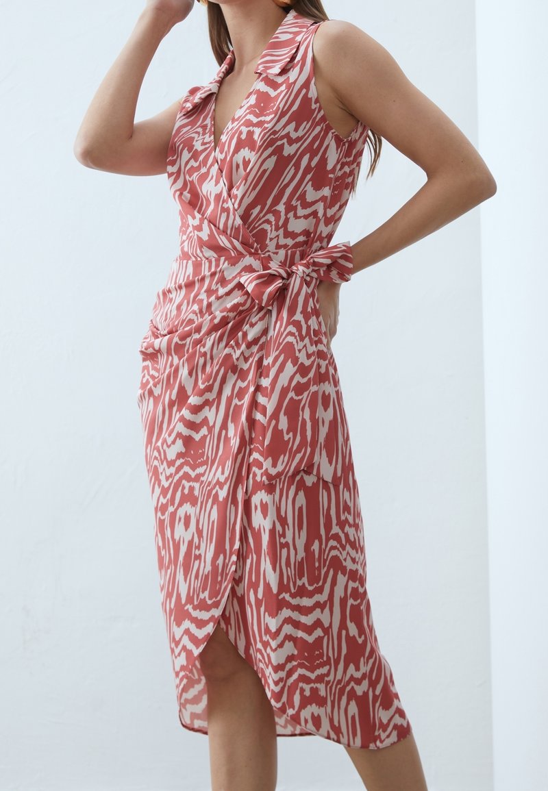 Femme portant une robe portefeuille sans manches à motifs corail et blanc avec col, nouée à la taille, debout devant un fond blanc uni.