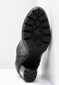 Bota de piel negra con un tacón cuadrado y una suela de goma negra texturizada que presenta patrones de dibujo definidos y acentos geométricos.