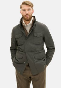 Giacca verde scuro realizzata in un tessuto liscio, con chiusura con zip, due tasche sul petto e design con colletto. Indossata sopra un maglione chiaro.