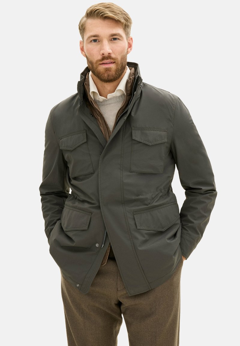Giacca verde scuro realizzata in un tessuto liscio, con chiusura con zip, due tasche sul petto e design con colletto. Indossata sopra un maglione chiaro.