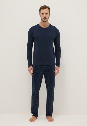 Man staat op blote voeten, draagt een marineblauw shirt met lange mouwen en een marineblauwe broek met kleine witte stippen tegen een lichte achtergrond.