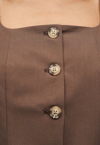 Καφέ ύφασμα με τρία μεγάλα στρογγυλά μπεζ κουμπιά τοποθετημένα κάθετα στη γραμμή λαιμού ενός ενδύματος, που δείχνει δέρμα από πάνω.