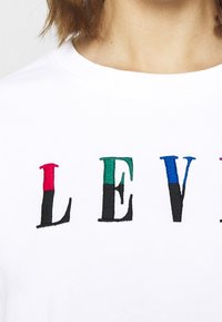 Gros plan sur l'encolure d'un t-shirt blanc brodée de lettres multicolores partielles "L", "E" et "V" en rouge, vert et bleu.