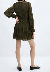 Oliivinvihreä, hulmuhameinen mekko, jossa on pitkähihaiset hihat, vyötärö on kapeaksi sommiteltu ja helma levenee. Mekossa on takana napitus ja kuvioitu kangas.