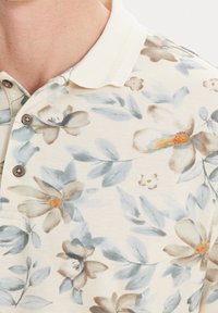 Květinová polo košile s krémovým pozadím, na kterém jsou světle modré listy a hnědé květy. Texturovaná tkanina se třemi knoflíky a pleteným límcem.