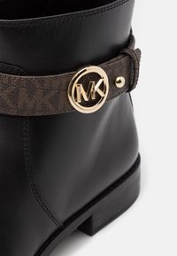 Botte de cheville en cuir noir avec une sangle à motif marron et un logo circulaire doré, dotée d'une semelle plate. Texture lisse avec des détails cousus.