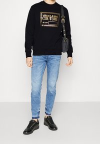 Svart sweatshirt med guld-logotyp, ljusblå denimjeans och svarta sneakers med guldaccenter. Avslappnad stil, minimal textur och detalj.