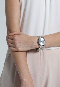 Zilveren metalen horloge met een minimalistisch rond wijzerplaat, lichtreflecterende schaal en een schakelarmband. Gekenmerkt op een pols, gecombineerd met een lichtblouse.