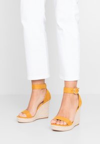 Žluté semišové sandály na klínu s páskem kolem kotníku, espadrilkový styl s pleteným jute klínem, otevřená špička a hladký texturovaný povrch.