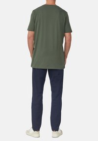 Oliwkowa koszulka z krótkim rękawem o luźnym kroju, w zestawie z ciemnonawigacyjnymi spodniami i jasnym obuwiem, stojąca na jednolitym tle.