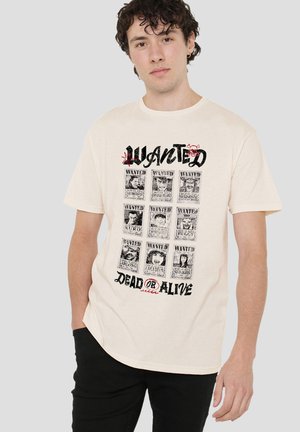 Young man with curly hair wearing a beige T-shirt featuring nine vintage-style "Wanted" posters and the text "Dead or Alive."