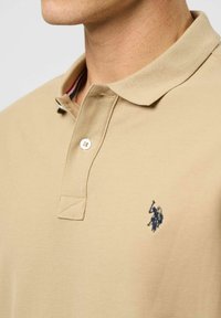 Detailní záběr na osobu, která má na sobě béžové polo tričko s límečkem, dvěma knoflíky a malým tmavomodrým vyšívaným logem na hrudi.