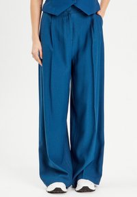 Pantalon large bleu en tissu lisse, avec des plis et une taille haute, associé à des baskets blanches, posé devant un fond neutre.
