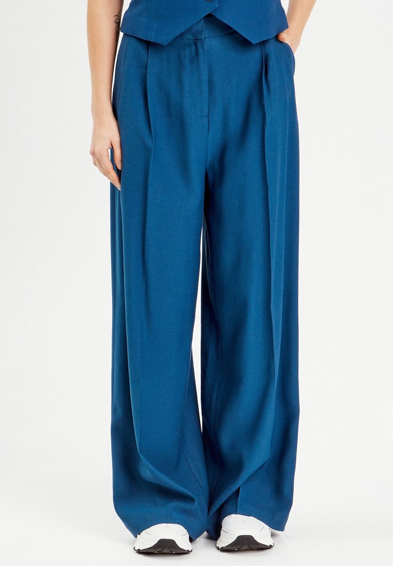 Pantalon large bleu en tissu lisse, avec des plis et une taille haute, associé à des baskets blanches, posé devant un fond neutre.