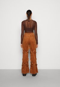 Bruine ribfluwelen flare pants met uitsparingen en decoratieve hardware. Doorzichtige lange mouwen top met horizontaal gestreept patroon.