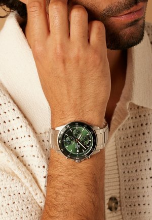 Zilveren metalen chronograafhorloge met een groene wijzerplaat, witte uurmarkeringen en een roestvrijstalen band, gedragen om een pols. Structuurrijke gebreide achtergrond.