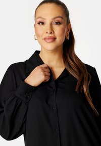 Blusa nera con colletto, apertura frontale con bottoni e maniche lunghe a sbuffo. Tessuto liscio e leggero con una consistenza morbida.