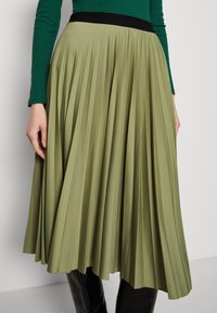 Falda midi plisada en verde oliva con cinturilla negra, con pliegues verticales y una textura suave, estilizada con una blusa ajustada.