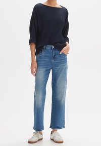 Marinblå vadderad stickad topp med lös passform, tillsammans med ljusblå högmidjade raka jeans och vita sneakers med bruna sulor.