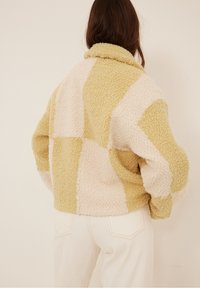 Dzianinowa kurtka z fakturowanego materiału w patchworkowym wzorze z beżowymi i jasnosłomkowymi sekcjami. Posiada złożony kołnierz i luźny krój.