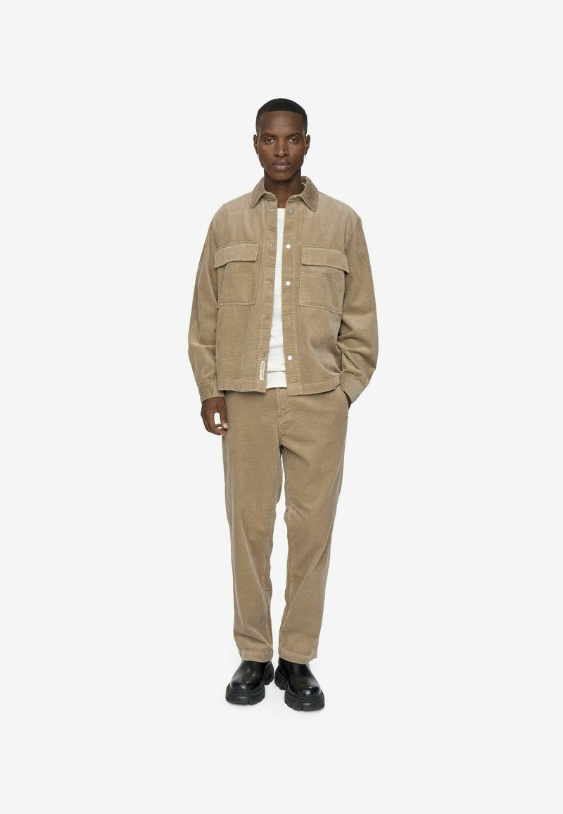 Brun kordfløjljakke med to frontlommer, parret med matchende kordfløjl bukser. Sorte sko fuldender outfittet. Enkel, afslappet design.
