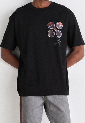 Black short-sleeve t-shirt with abstract floral design and text on chest, worn with light gray jeans featuring a side stripe.