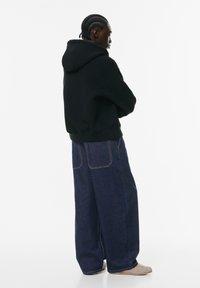 Zwarte hooded sweatshirt gecombineerd metwijdpijpzwarte denim jeans. Kenmerkt zichtbare stiksels en een relaxte pasvorm. Schoenen zijn licht van kleur en minimalistisch.