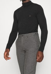 Schwarzer Rollkragenpullover aus glattem Material mit einem dezenten Logo. Kombiniert mit gemusterten grauen Hosen, die eine taillierte Passform haben.